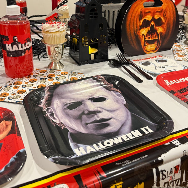A Halloween-themed party table setup featuring Michael Myers printed paper plates, a black haunted house sign, red drink bottles, red balloons, and spooky decorations.