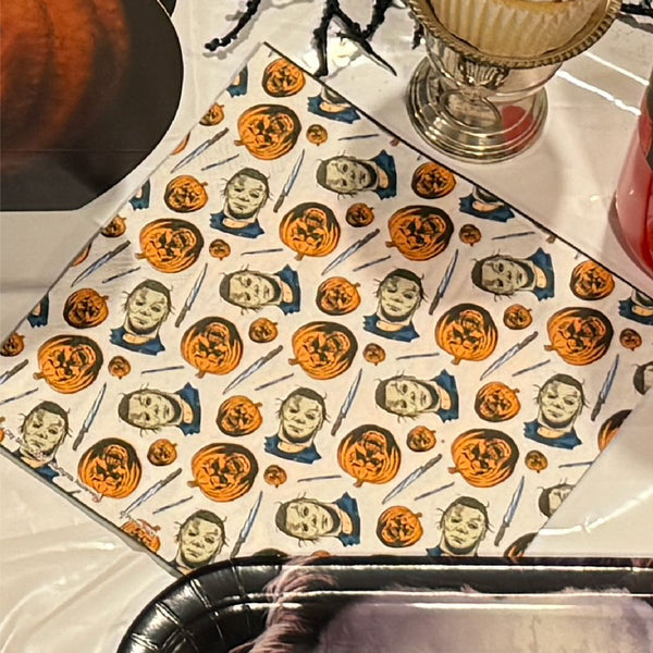 A Halloween-themed paper napkin featuring a repeating pattern of jack-o'-lantern pumpkins and ghostly faces on a white background, styled on a table with a cupcake, a pumpkin, and black spider decorations.