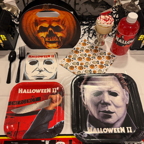 Halloween-themed party tableware set featuring Michael Myers prints on square paper plates, a red drink bottle labeled HALLOWEEN, patterned napkins, a plastic fork, and black plastic cutlery on a white tablecloth.