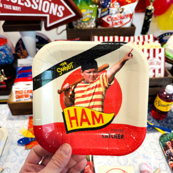 Hand holding a tray with a vintage-style 'Ham' label design, surrounded by arcade game paraphernalia.