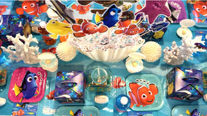 Overhead view of a Finding Nemo party table with a clamshell centerpiece, white coral and faux sea-urchin accents, pearl-in-shell decorations, character standees of Nemo, Dory, Marlin and Bruce, and square Dory and Nemo plates on a blue ocean table cover.