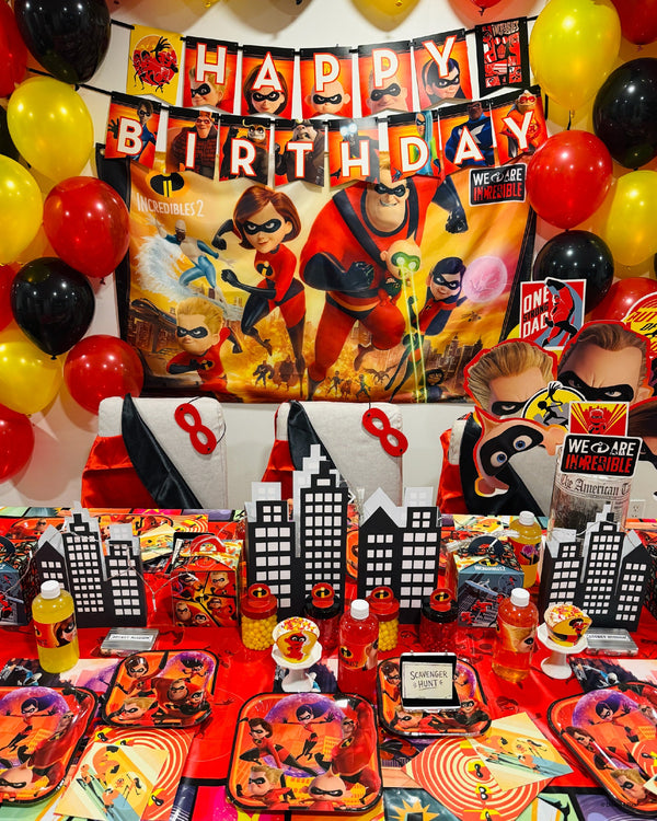 Incredibles 2 birthday party table set with themed plates, favor boxes, candy jars and a black paper city skyline