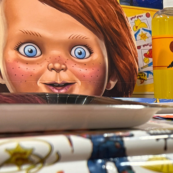 A Chucky themed party setup featuring a white tablecloth printed with Chucky illustrations and text, along with matching paper cups, cups, and a large Chucky face cutout in the background.
