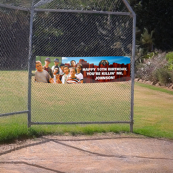 Birthday banner with a group of people displayed on a chain-link fence in an outdoor setting. Sandlot-themed birthday banner attached to a baseball field fence with kids and dog design and “You’re Killin’ Me, Johnson” text.